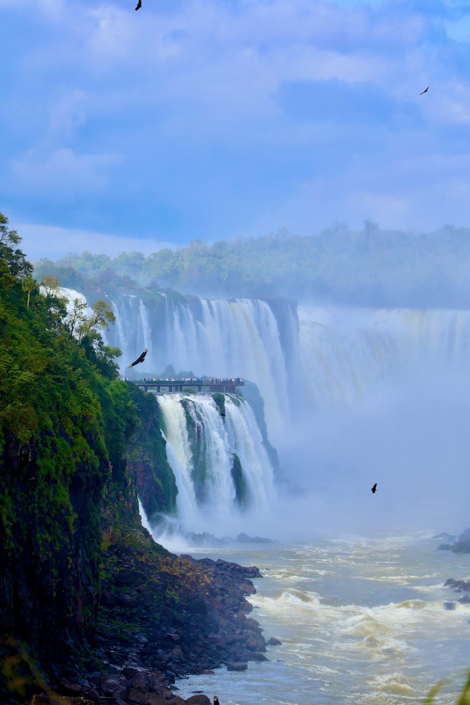 Visitors stand on a viewing platform overlooking massive cascading waterfalls.